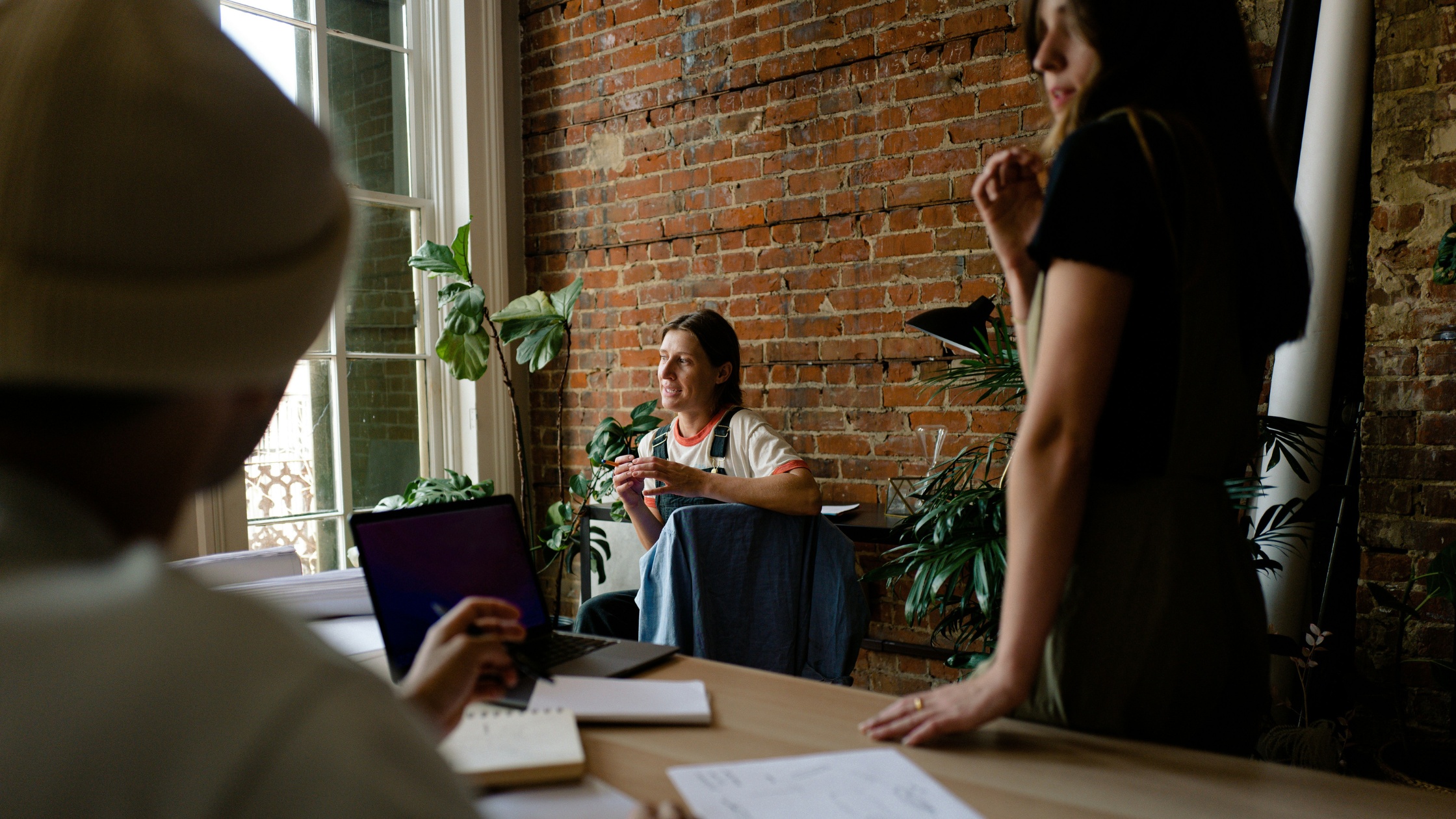 Three coworkers in a casual meeting, discussing ideas around a table in a modern office with brick walls and plants.